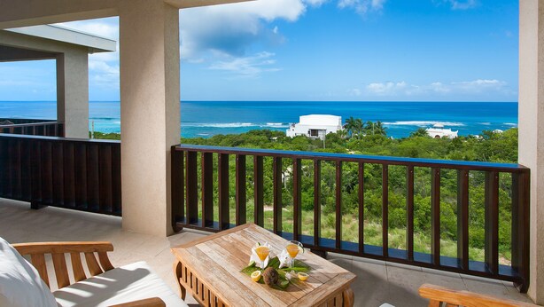 A balcony with a wooden table, two chairs, and a view of the ocean and greenery.