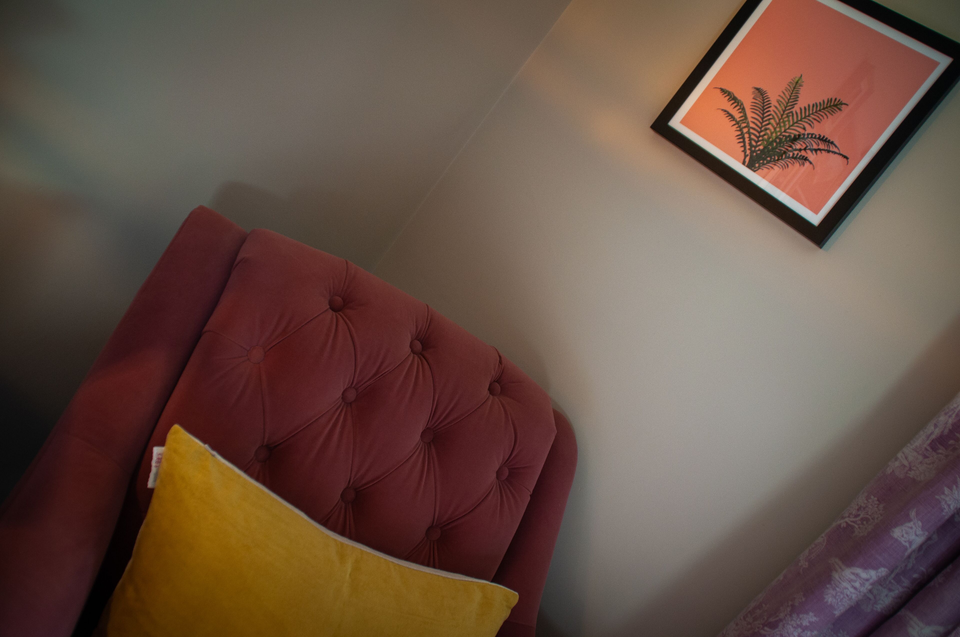 A tufted red headboard with a yellow pillow, a framed plant on the wall, and a purple curtain.