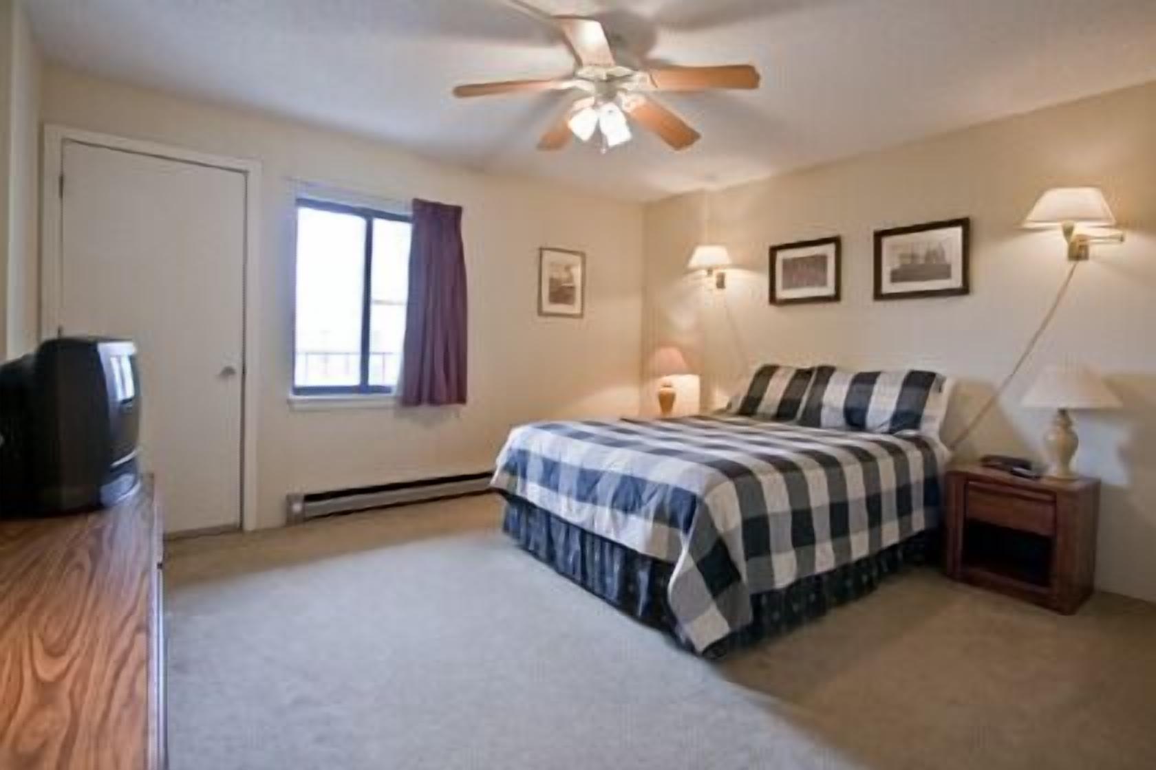 A bedroom with a bed, a ceiling fan, a window with curtains, a nightstand with a lamp, and a television on a wooden dresser.