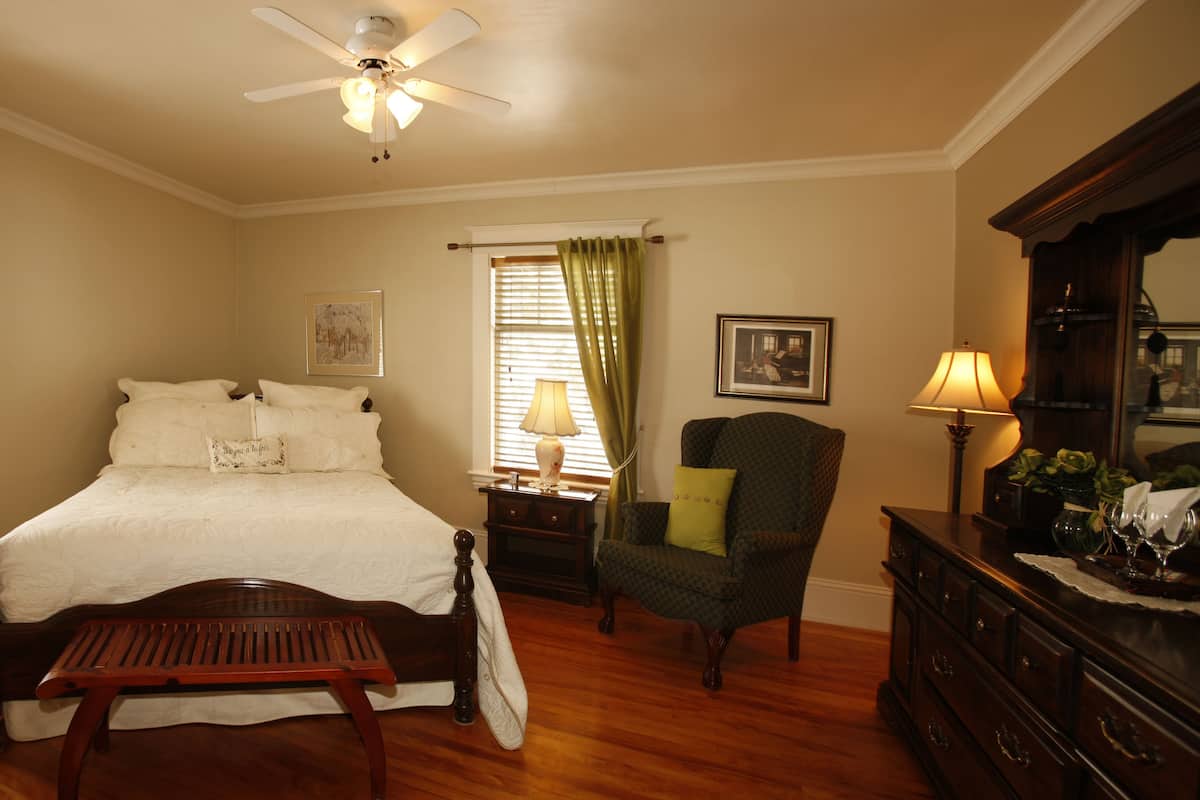 A bedroom with a bed, a chair, a dresser, a nightstand, a lamp, a window with curtains, a ceiling fan, and a radiator.