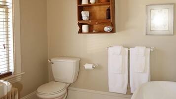 A bathroom with a toilet, a wooden shelf with kitchen items, a radiator, and a bathtub.