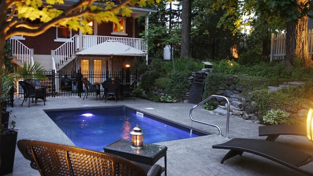 A swimming pool with lounge chairs and a table, surrounded by a stone wall and greenery. - B&B Au Virage (Magog)