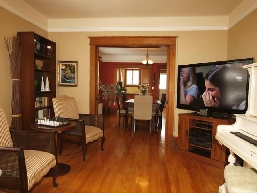 A living room with a wooden floor, a piano, a television, and a dining area in the background.