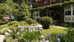 A brick building with ivy-covered walls, a well-maintained garden with purple flowers, and a sign indicating a business. - B&B Au Virage (Magog)