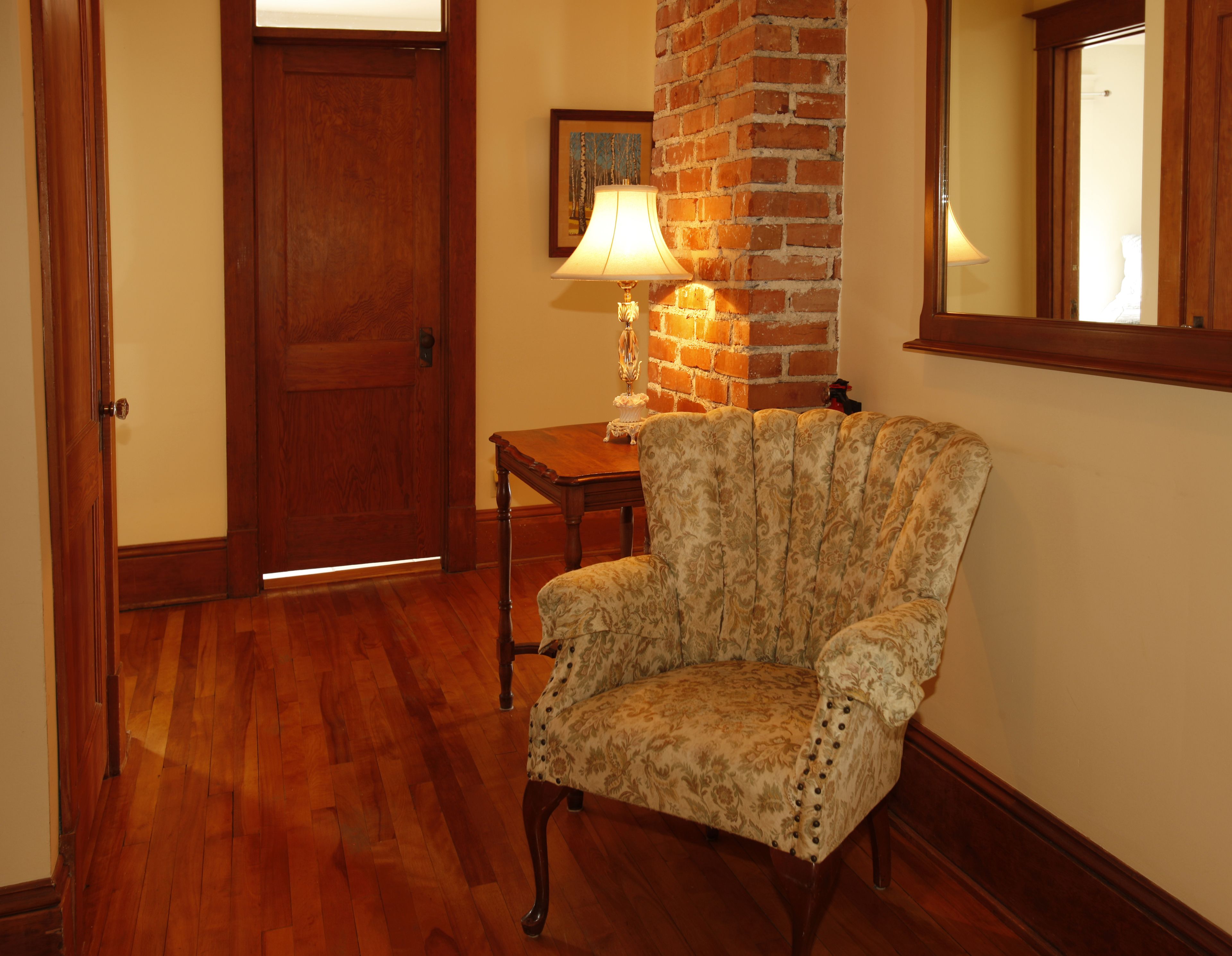 A wooden chair with a floral pattern, a wooden table with a lamp, and a brick wall.