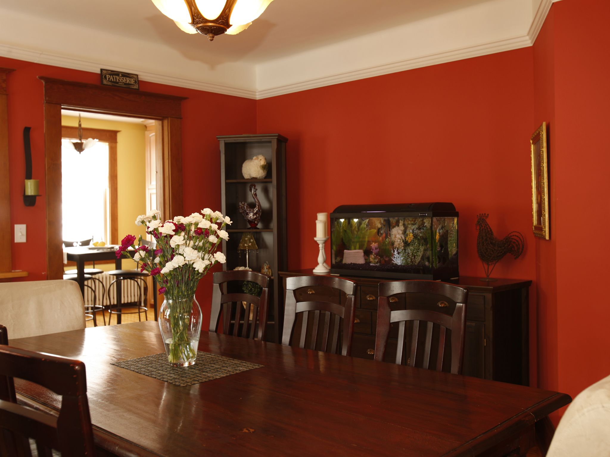 A dining room with a wooden table, chairs, a vase of flowers, a fish tank, and a red wall.