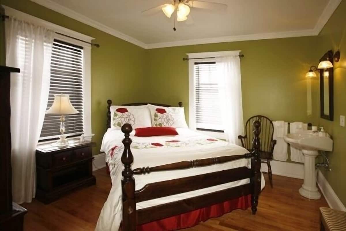 A bedroom with a wooden bed, a nightstand, a chair, a window with blinds, a ceiling fan, and a white sink.