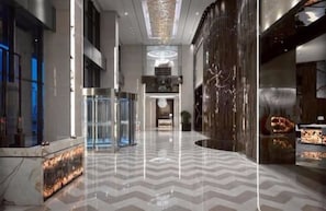 A spacious lobby with a modern design, featuring a marble floor, a glass elevator, and a central decorative light fixture.