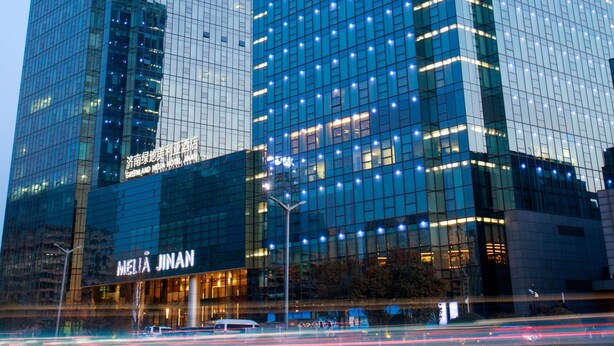 Um edifício alto com janelas de vidro refletivas e placas com os dizeres 'GREENLAND GROUP' e 'MELIA JINAN'.