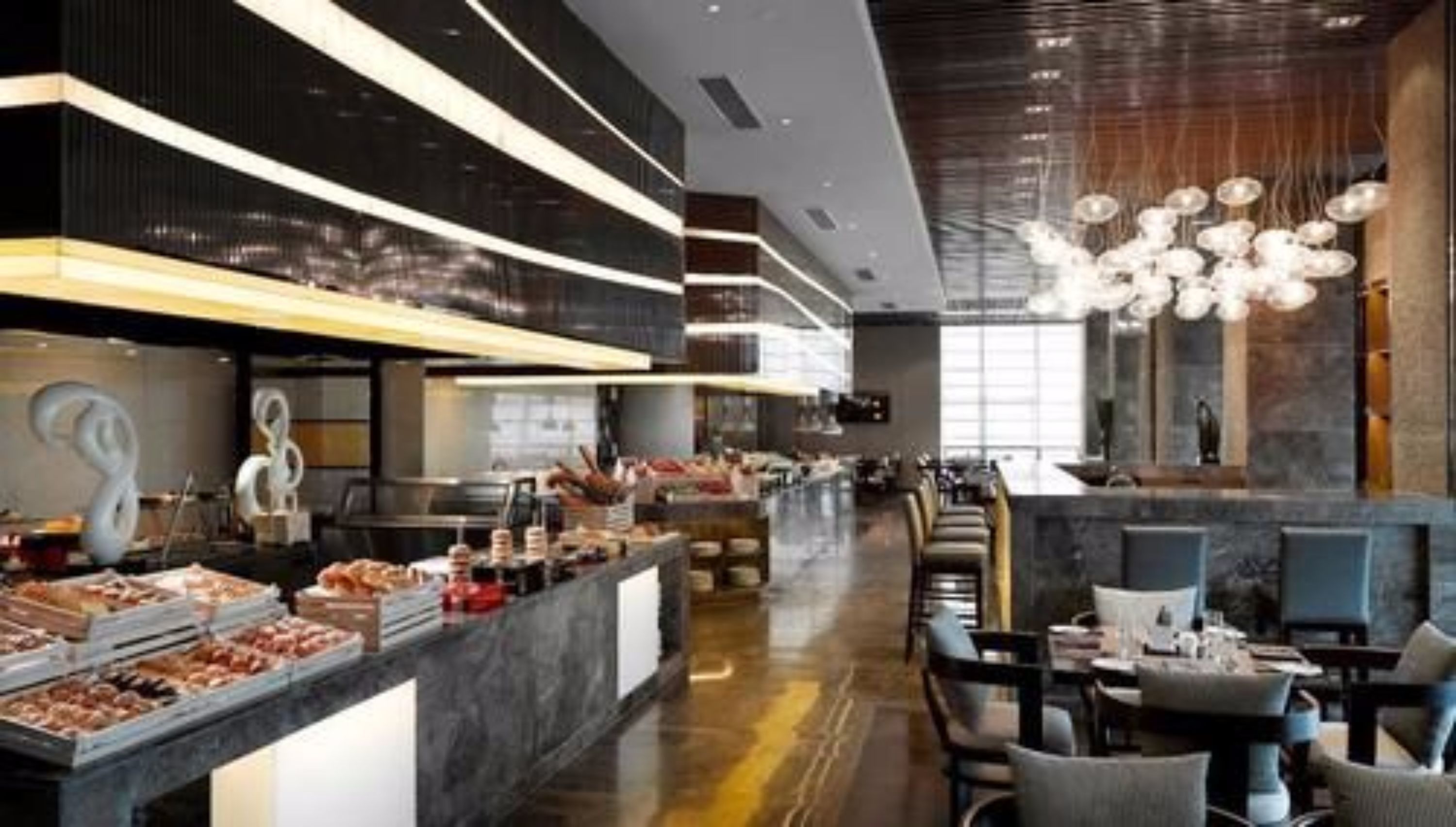 Un ristorante moderno con un lungo bancone che espone diversi prodotti alimentari, zona con tavoli e sedie e un lampadario decorativo.