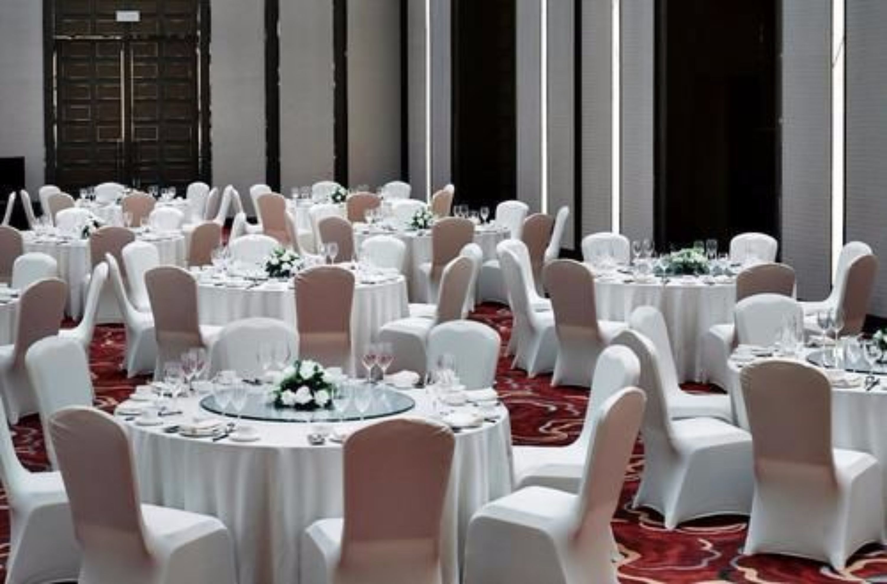 Une salle de banquet avec des tables rondes dressées pour le repas, des chaises blanches et une moquette rouge.