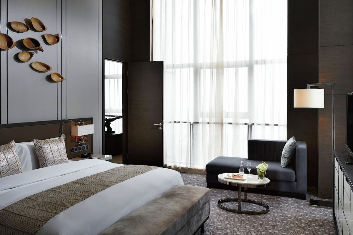 A hotel room with a bed, a sofa, a small round table, a TV, and a large window with sheer curtains.