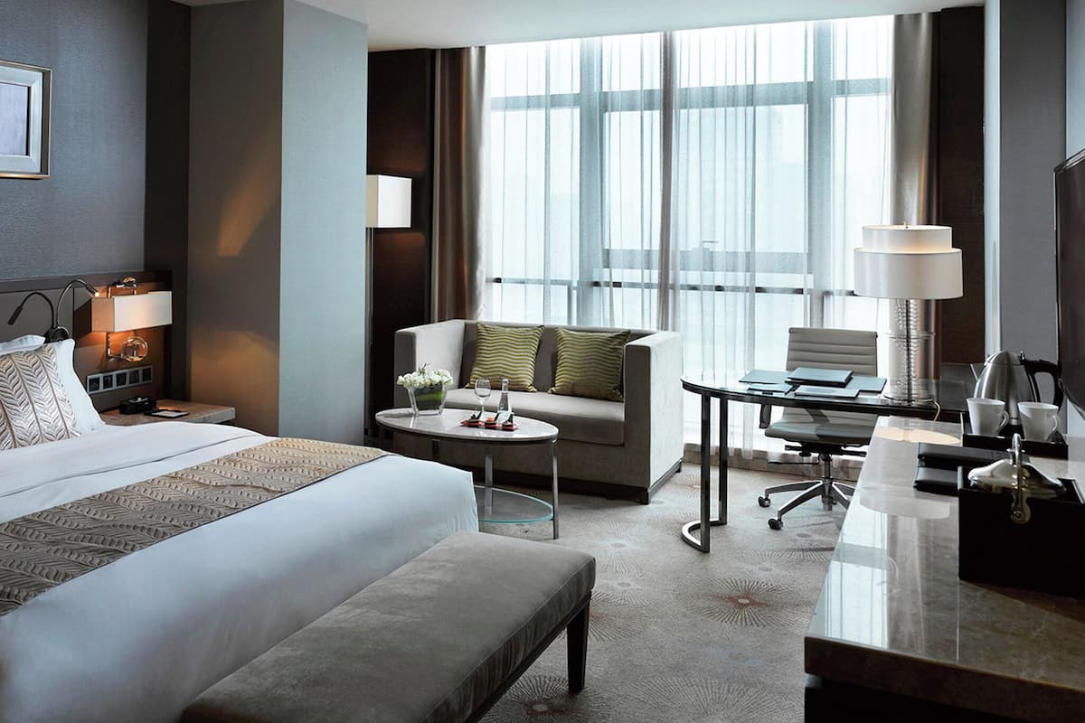 A hotel room with a large bed, a sitting area with a sofa and armchair, a desk with a chair, and a large window with curtains.