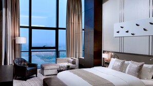 Quarto de hotel moderno com uma cama grande, área de estar com sofá e poltronas, uma mesinha e vista para a cidade através de grandes janelas.