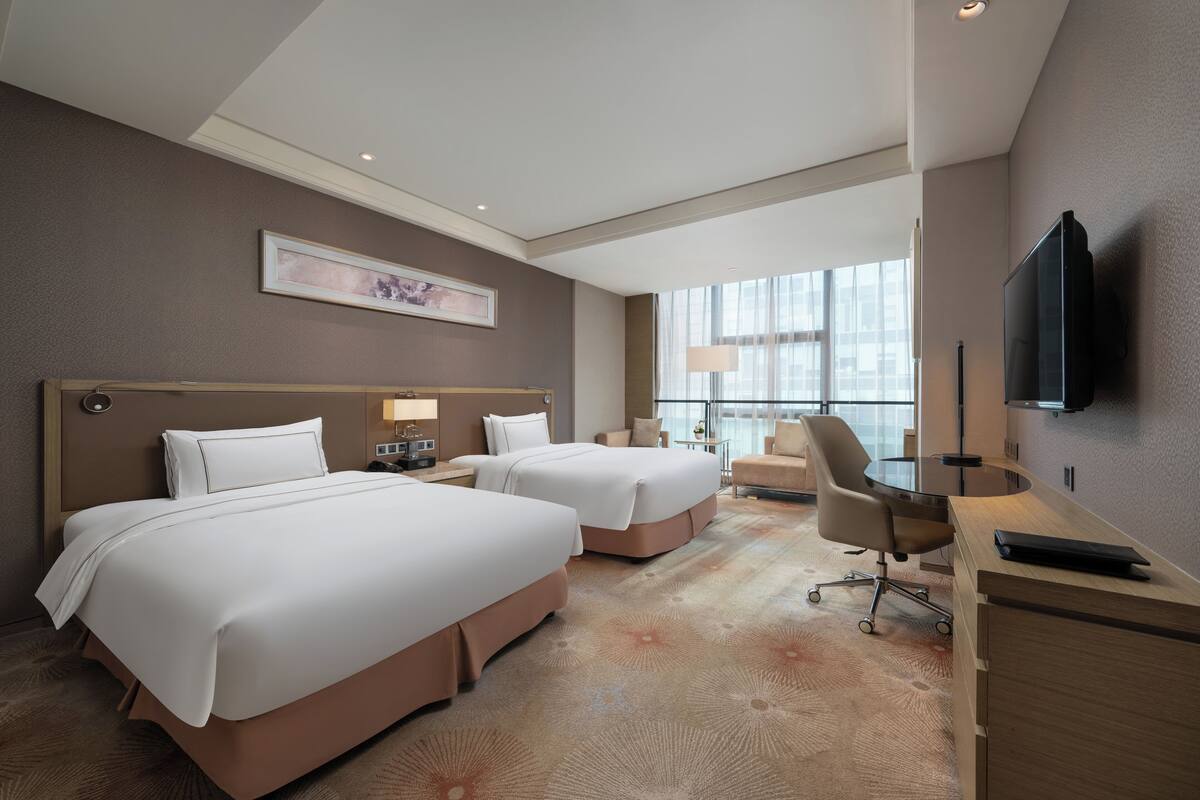 A hotel room with two beds, a desk, a chair, a TV, and a large window.