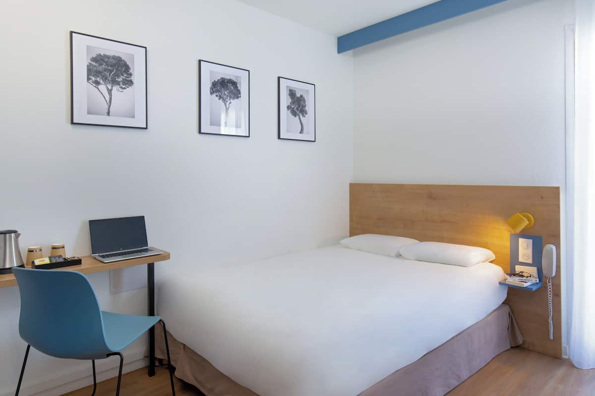 Habitación de hotel con cama, escritorio con una computadora portátil, silla y mesita de noche con teléfono y lámpara.