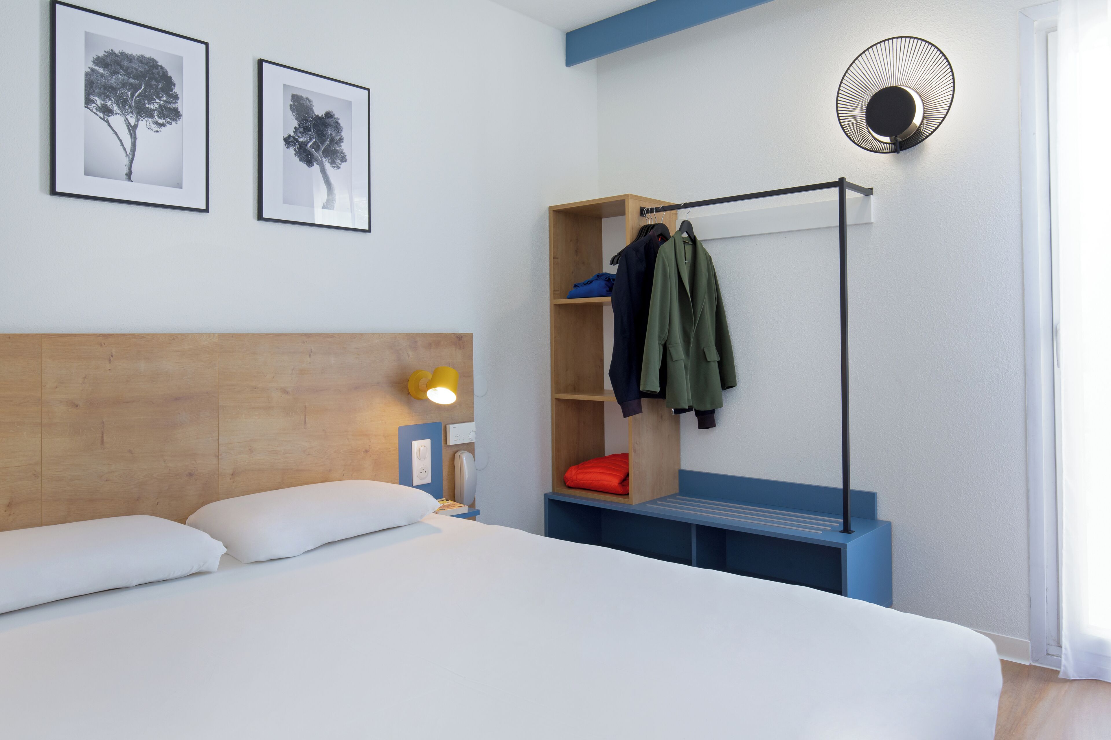 Una habitación de hotel con una cama, un cabecero de madera, un perchero con ropa y dos cuadros en la pared.