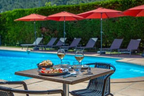 Una mesa junto a la piscina con un cesto de pan, un plato de comida y dos copas de vino, con sillones de descanso y sombrillas rojas al fondo.
