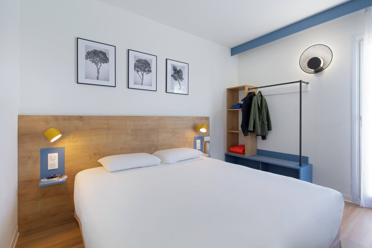 Un dormitorio con cabecera de madera, cama con sábanas blancas, mesita de noche con lámpara y estante en la pared con ropa.