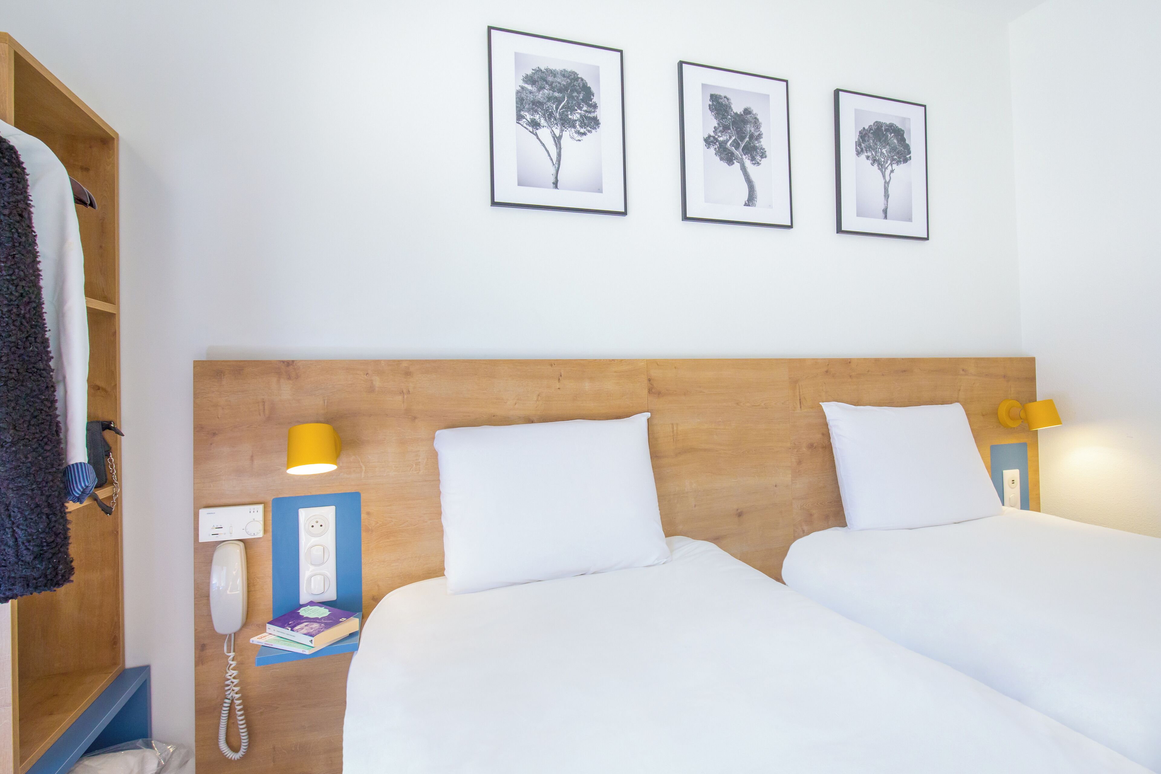 A hotel room with two beds, a wooden headboard, a bedside table with a lamp, a phone, and framed pictures on the wall.