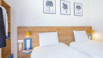 A hotel room with two beds, a wooden headboard, a bedside table with a lamp, a phone, and framed pictures on the wall.