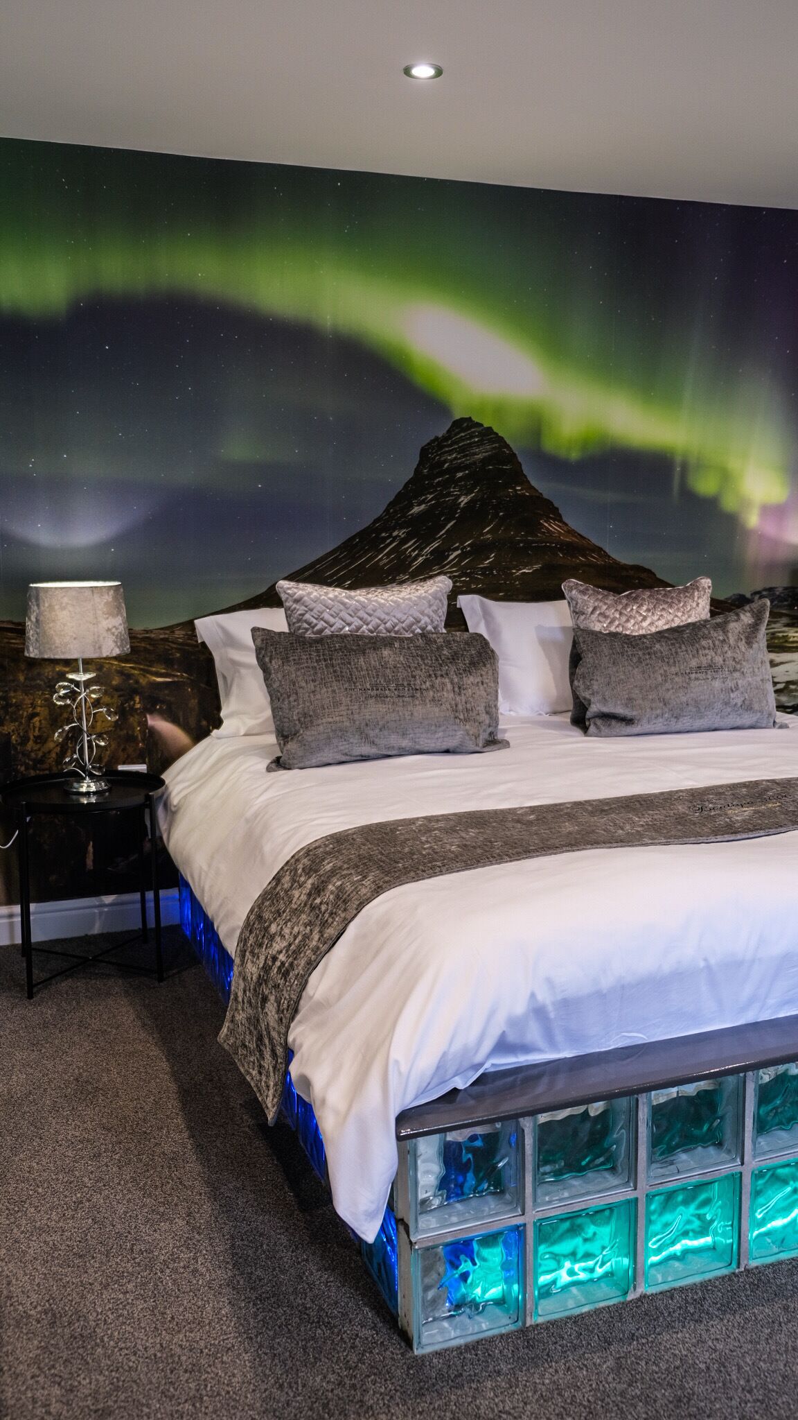 Ein Schlafzimmer mit Bett, Kissen, einem Nachttisch, einer Lampe und einer Wandmalerei von Bergen und Polarlichtern.