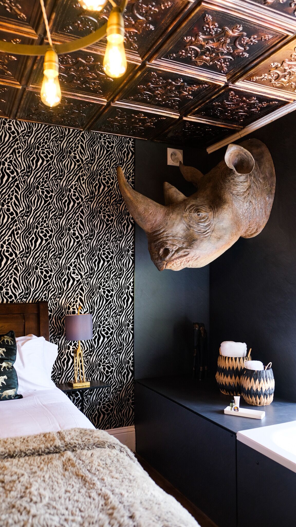 Une pièce avec un mur à motifs zébrés, une tête d’animal empaillée accrochée au mur, un lit, une table de chevet et une lampe.
