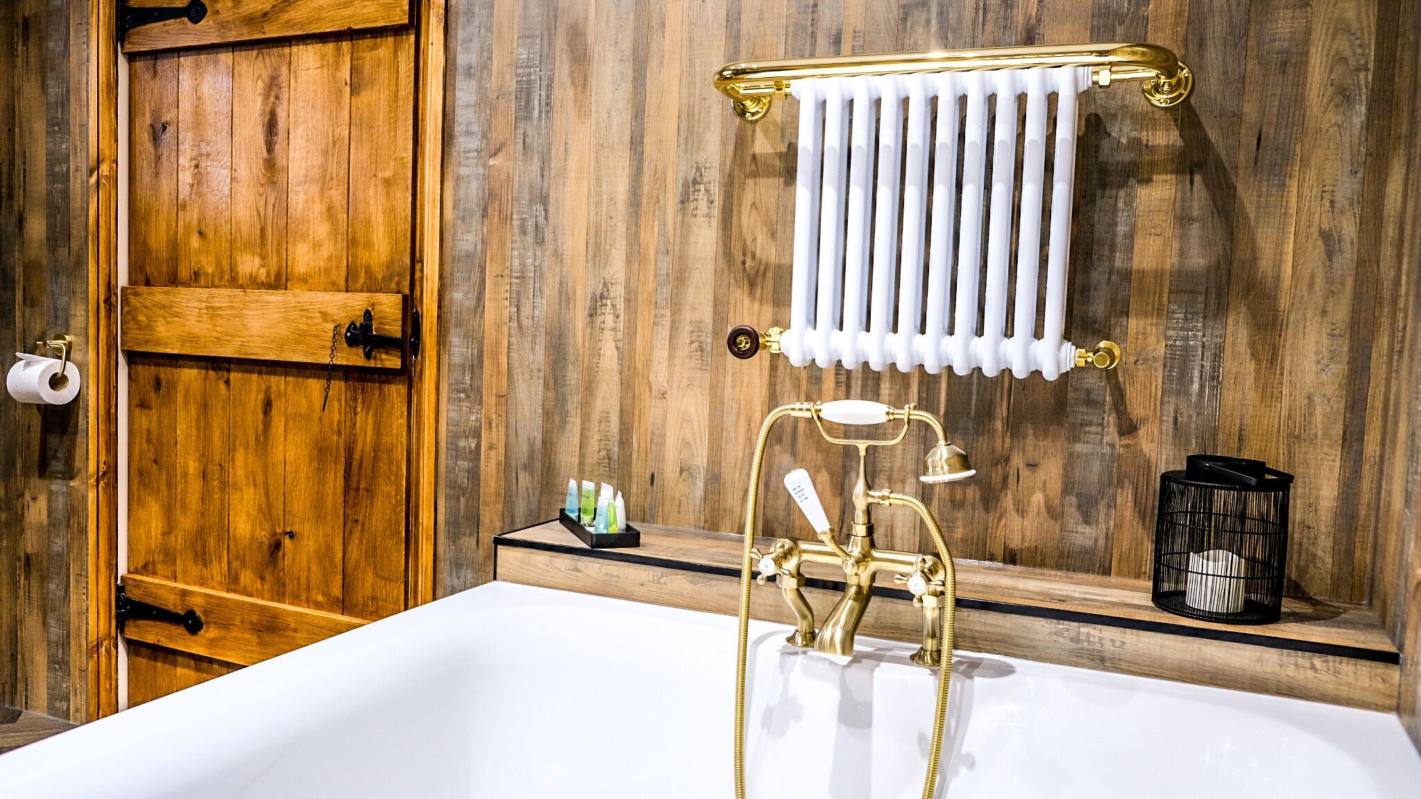 Een badkamer met een houten wand, een wit bad, een gouden kraan, een handdoekhouder, een toiletrolhouder, en een klein plankje met toiletartikelen.