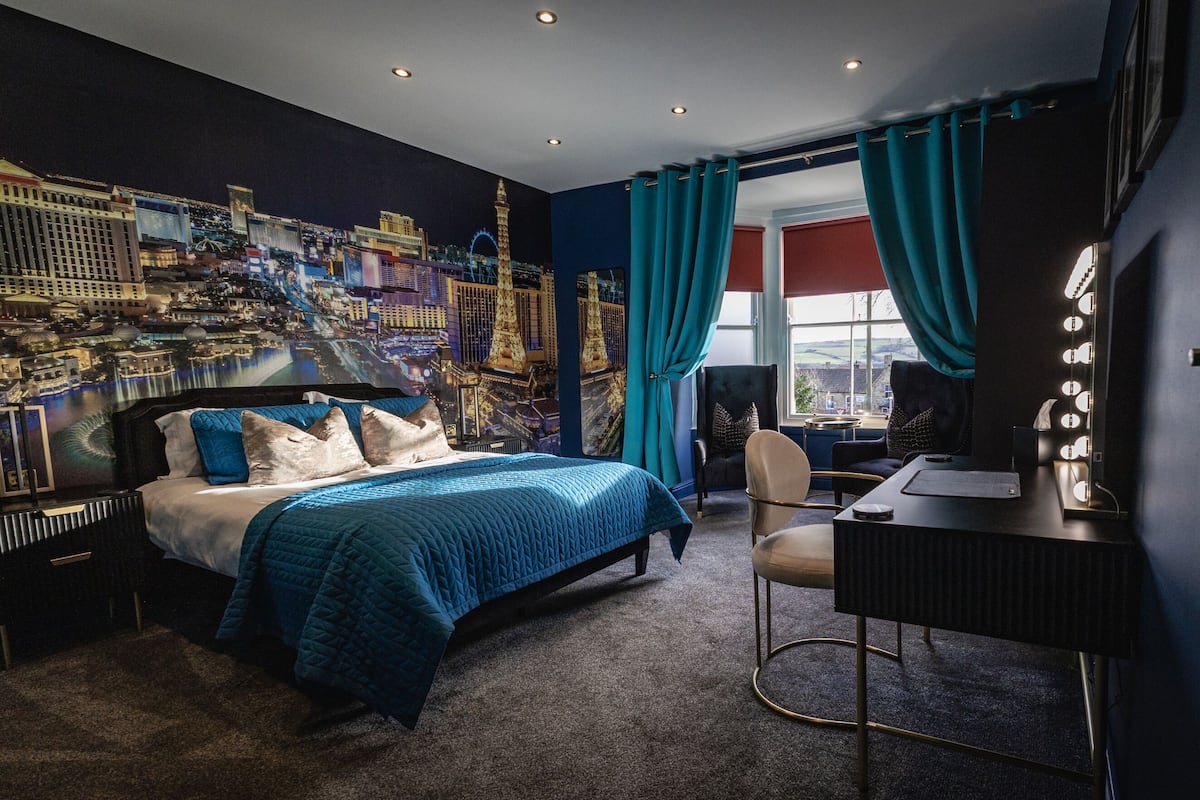 Una habitación de hotel con cama, un escritorio con silla, un ventanal con cortinas y un mural en la pared con vista a un paisaje urbano.