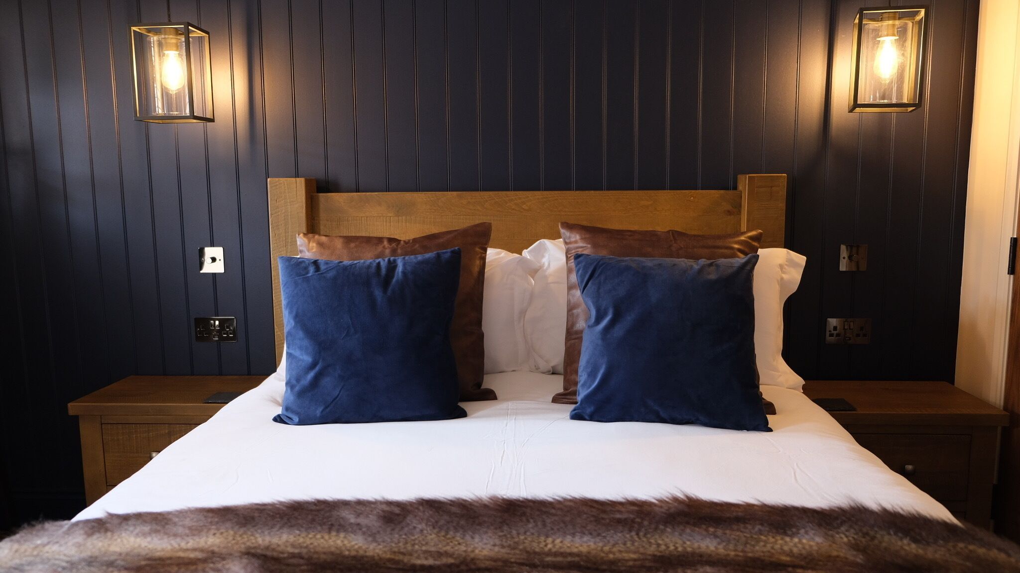 A hotel room with a wooden headboard, two blue pillows, a white bedspread, and two wall-mounted light fixtures.