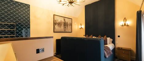 A compact hotel room with a bed, a small table, a chair, a wall-mounted picture, a chandelier, and a patterned wall.