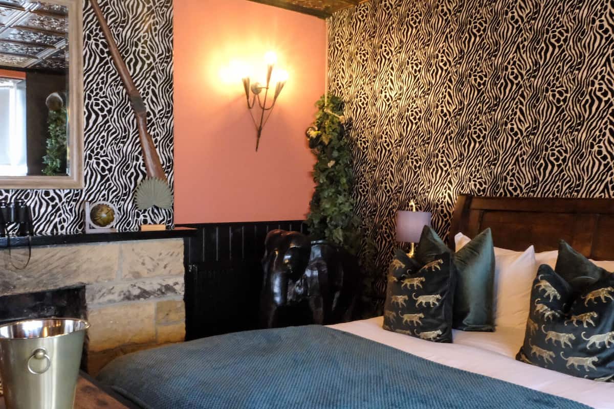 Un dormitorio con pared estampada de cebra, cama con almohadones con motivos animales, mesita de noche con lámpara y aplique de pared.