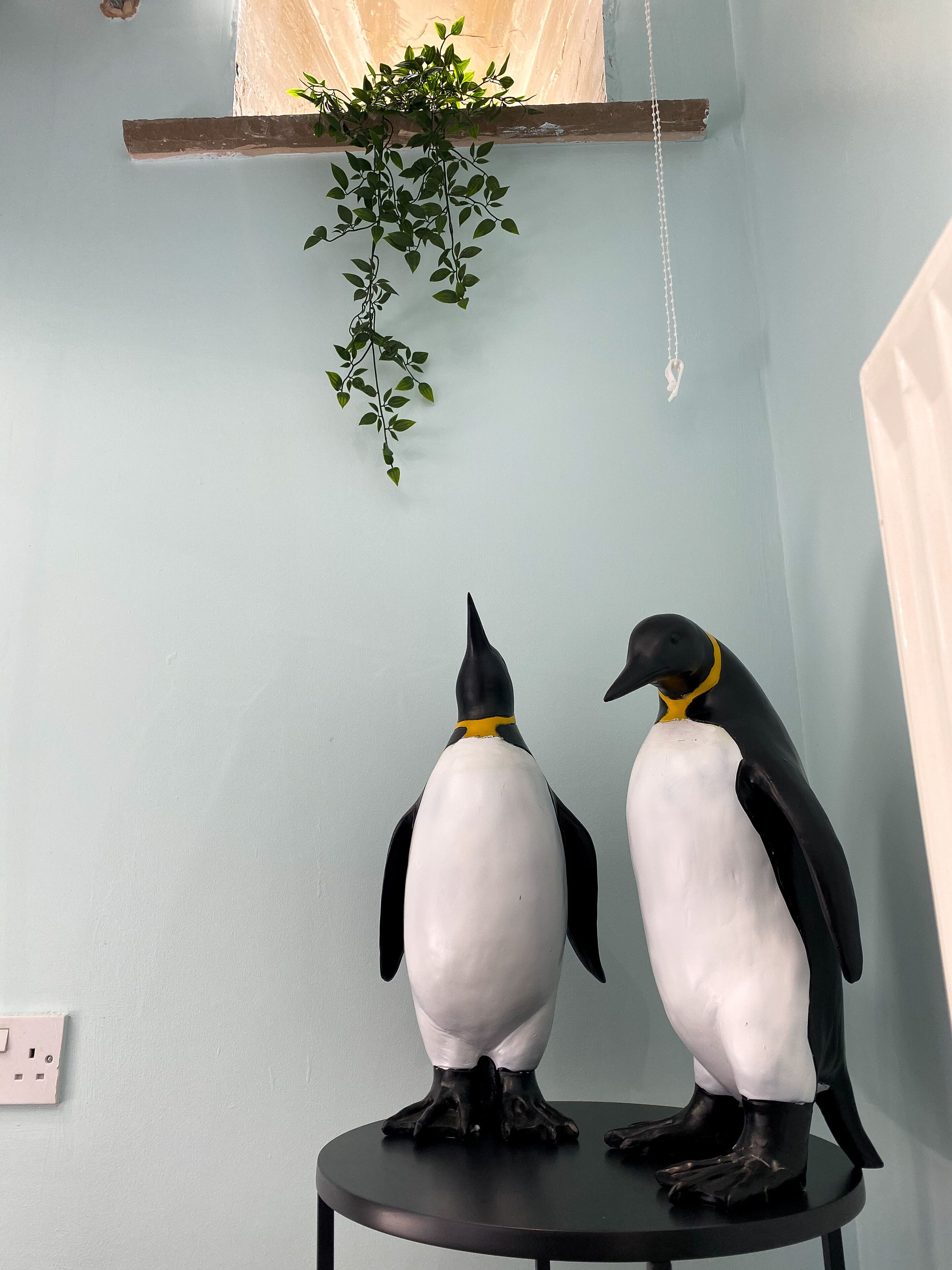Dos estatuas de pingüino sobre una mesa redonda negra frente a una pared azul claro.