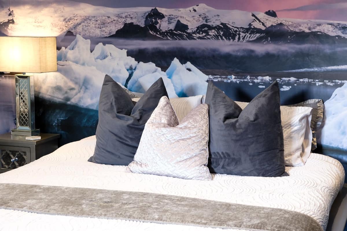 Una cama con colcha blanca y varias almohadas, una lámpara en una mesita de noche, y un mural grande con un paisaje montañoso nevado.