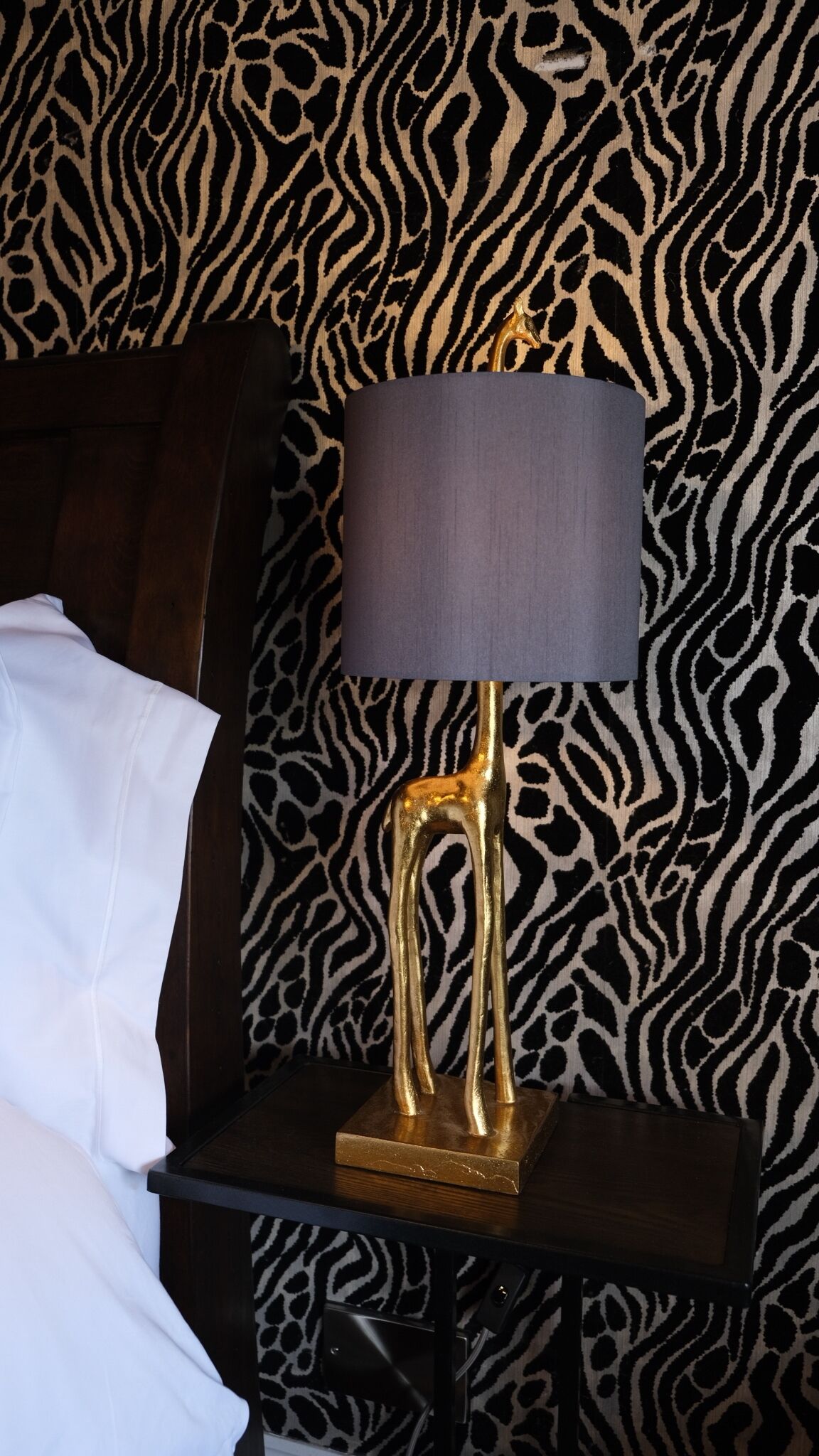 Una cama con sábanas blancas, un cabecero de madera oscura y una mesita de noche con una lámpara dorada y una pantalla negra.