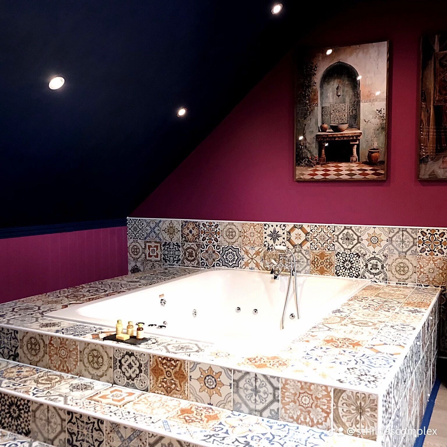 A bathroom with a large bathtub, patterned tiles, a red accent wall, and a framed artwork above the tub.