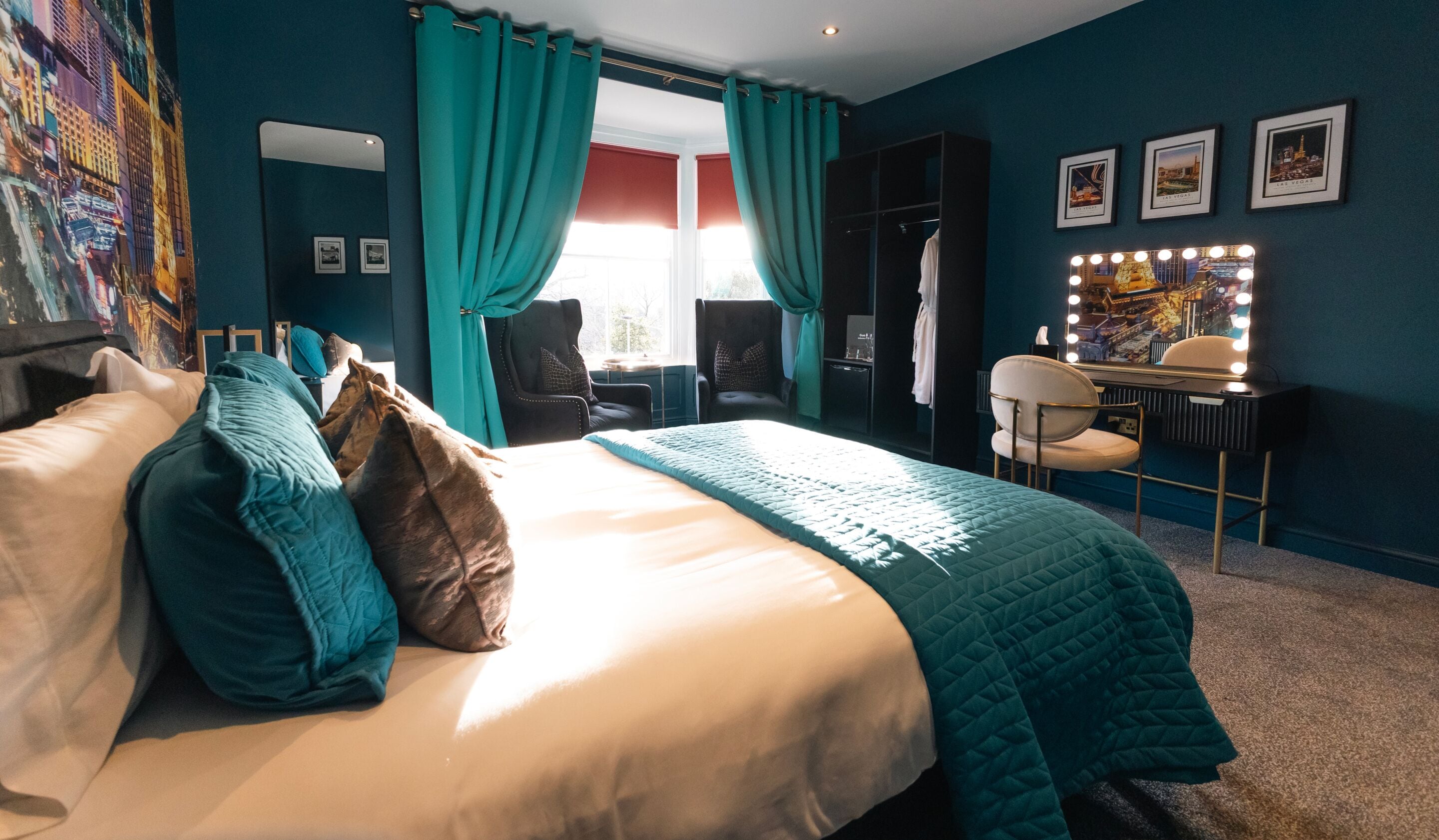 A hotel room with a large bed, teal curtains, a vanity with lights, and framed pictures on the wall.