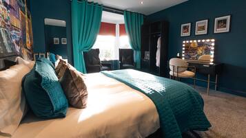 A hotel room with a large bed, teal curtains, a vanity with lights, and framed pictures on the wall.