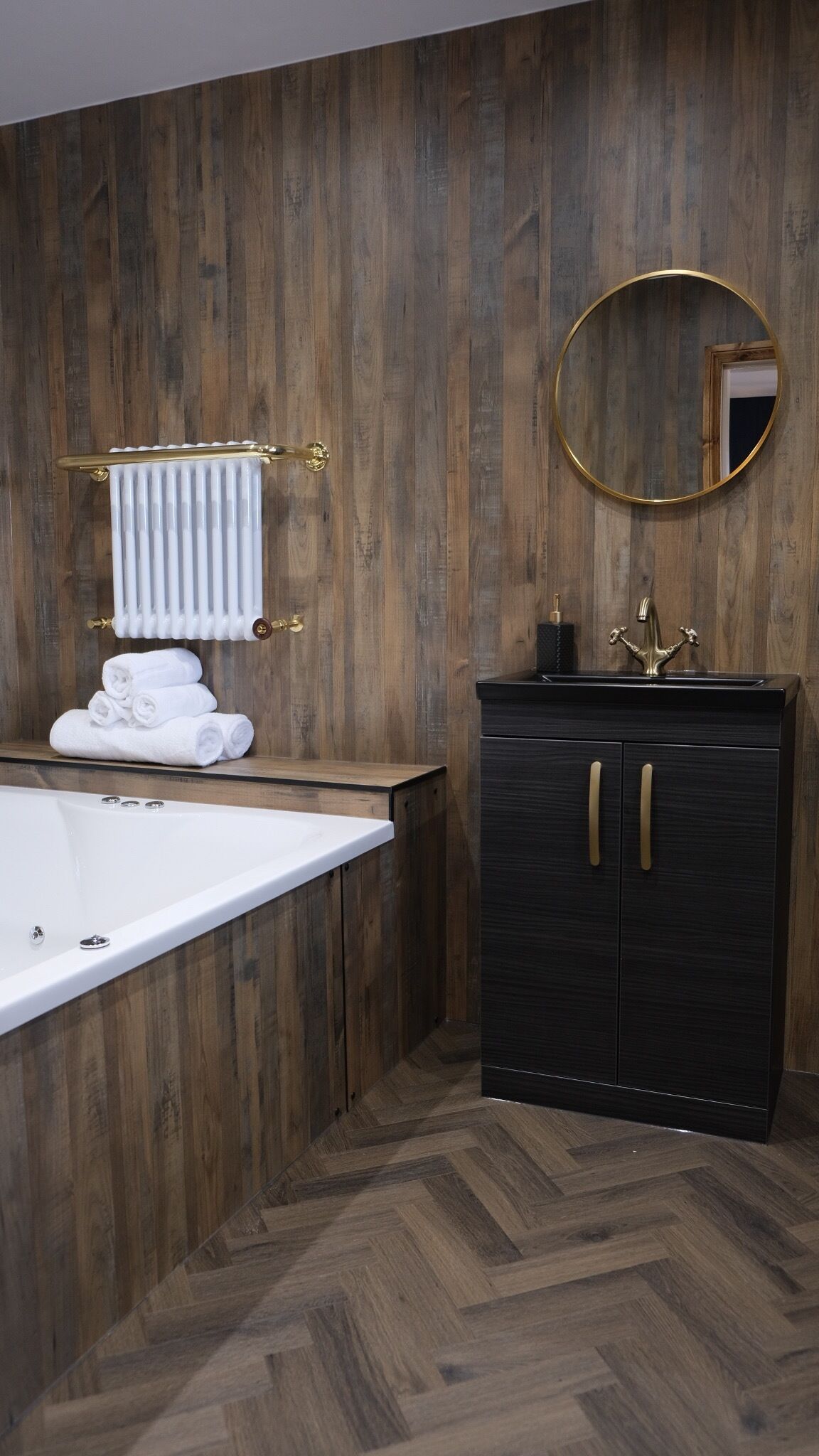 A modern bathroom with a wooden wall, a white bathtub, a black cabinet with gold handles, and a round mirror.
