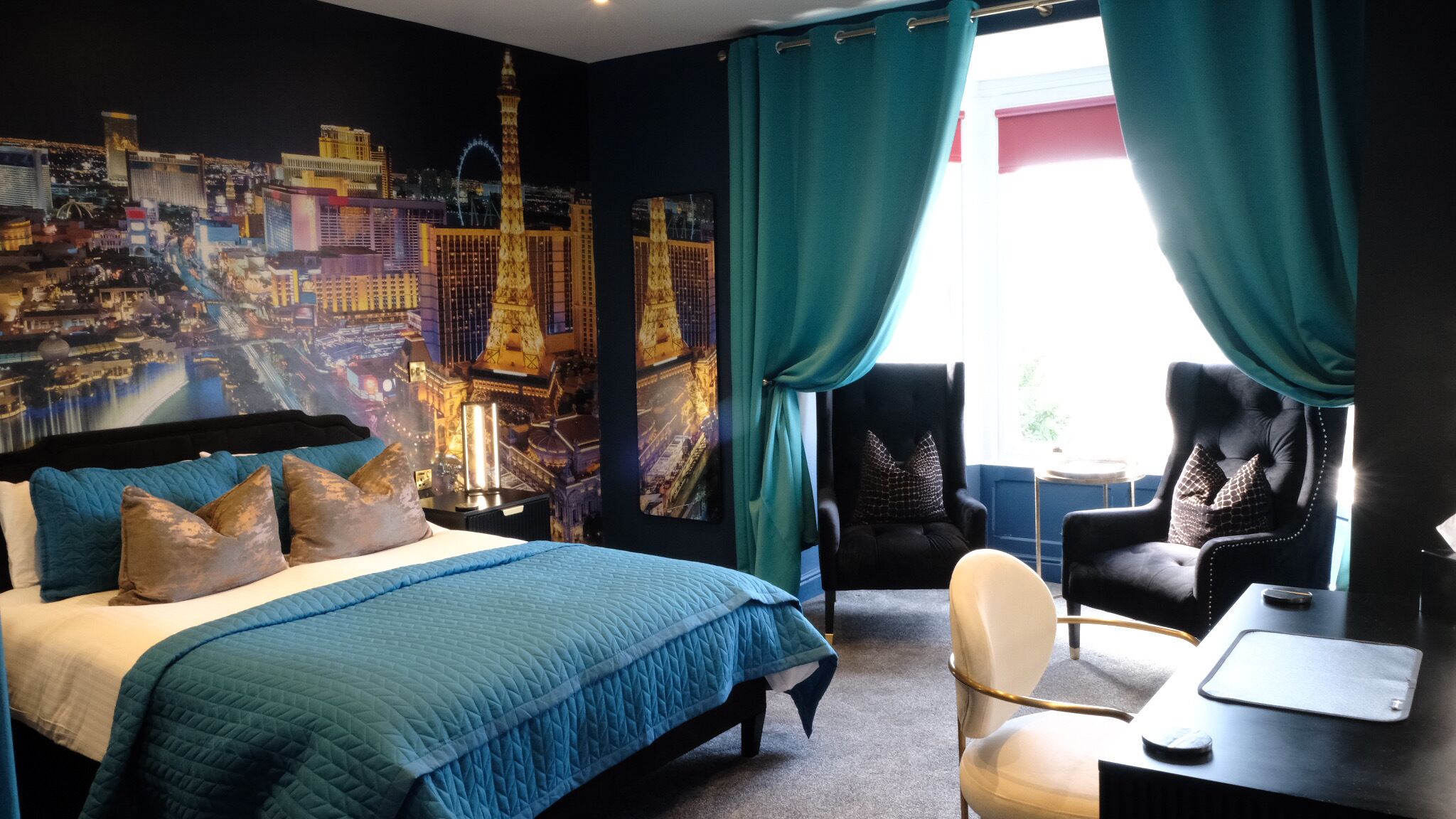 Une chambre d’hôtel avec un grand lit, deux fauteuils, un bureau et une fresque représentant un paysage urbain sur le mur.