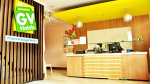 A hotel reception area with a wooden reception desk, a sign for 'GV Hotels', and a sign for 'Practical Budget Rooms'.