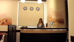 A reception desk with three clocks above it, a woman standing behind the desk, and a wall with a scenic mural. - Hotel Classic (Kharkiv)