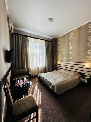 Relax in this inviting room with a comfortable bed, bedside table, and chair, surrounded by a decorative wallpaper pattern and a large window with sheer curtains.