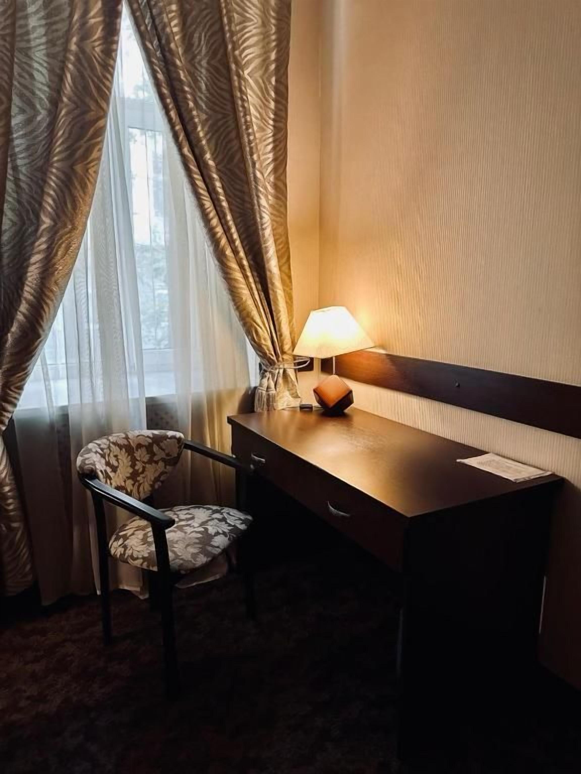 A hotel room with a desk, chair, lamp, and curtains.