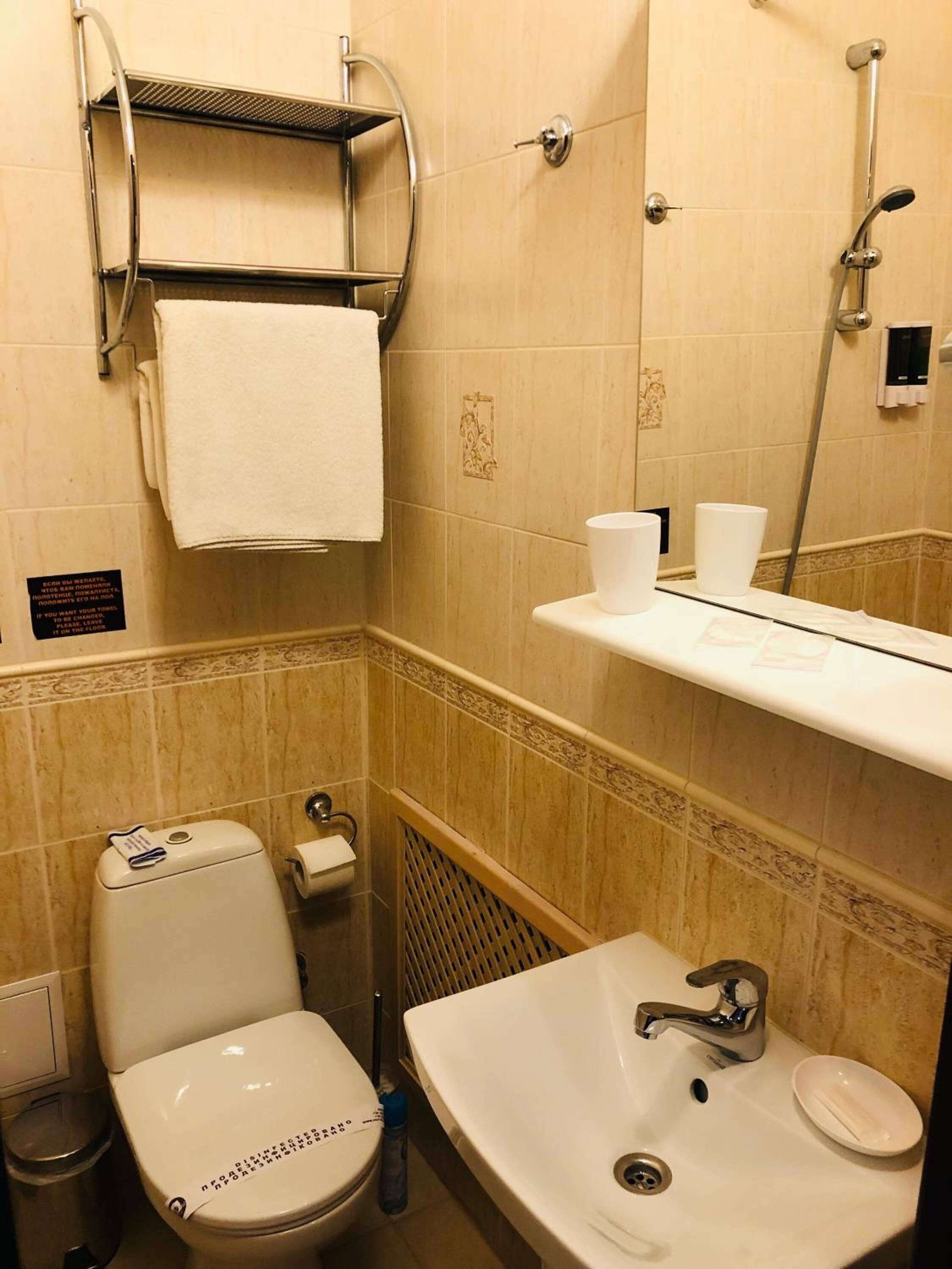 A small bathroom with a toilet, sink, and shower. There is a towel rack above the toilet and a shelf with cups.