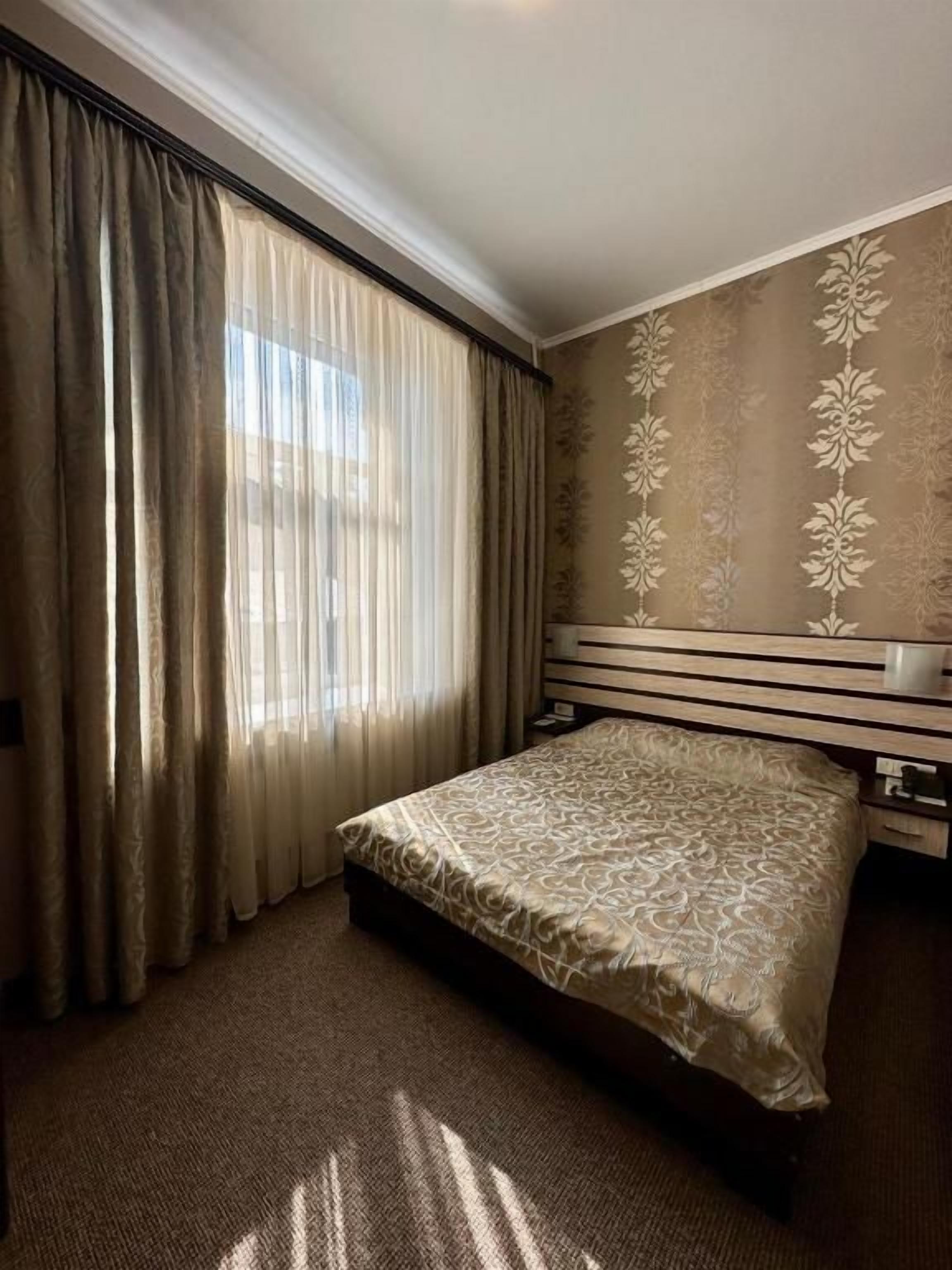 A hotel room with a bed, curtains, a patterned wall, and a carpeted floor.
