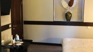 A hotel room with a bed, a desk with a chair, a small table with a vase, and a wall-mounted TV.