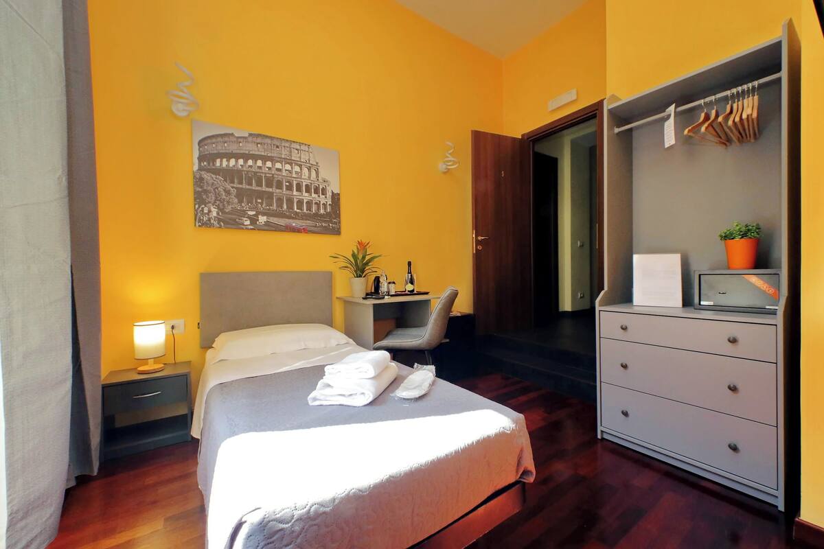 A hotel room with a bed, bedside table, lamp, chair, desk, wardrobe, and a painting of the Colosseum.