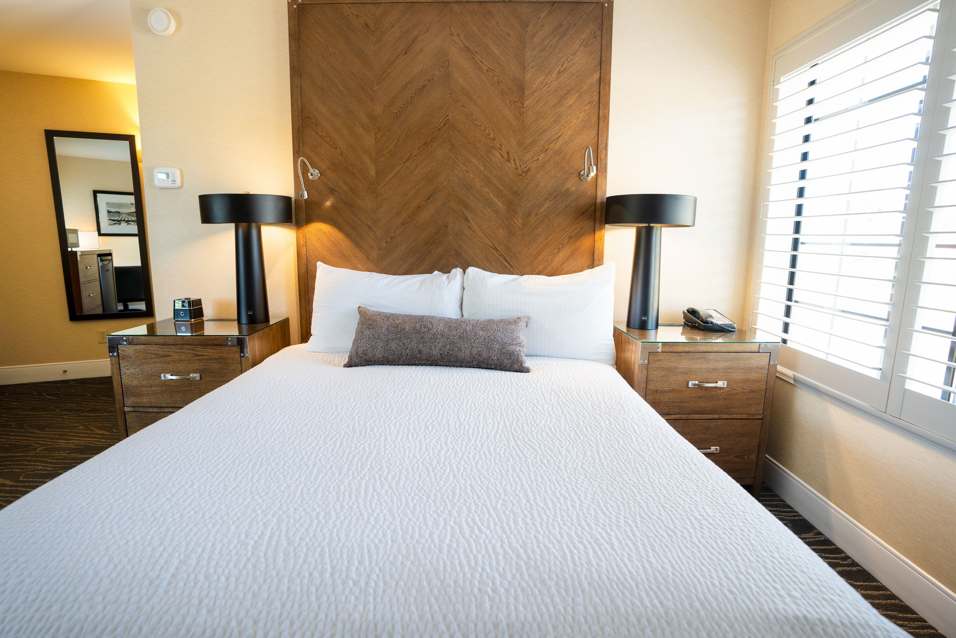 Une chambre d’hôtel avec un grand lit, deux tables de chevet, une commode, un miroir et une fenêtre avec des stores.