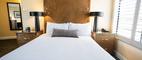 Une chambre d’hôtel avec un grand lit, deux tables de chevet, une commode, un miroir et une fenêtre avec des stores.
